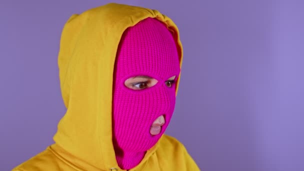 ピンクのバラクラバと黄色のフードで彼女の人生について紫色の背景に熟考の若い女性のクローズアップ 思慮深い女性でマスク離れて見て — ストック動画