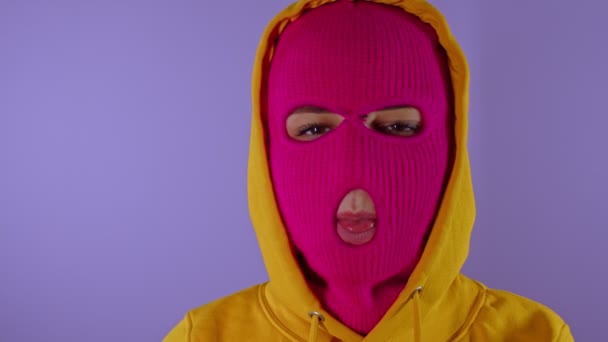 Close up of unrecognizable woman in pink balaclava shows her tongue on purple background. Unknown female in mask sticks out tongue and hides it, looking at camera. — Stock Video