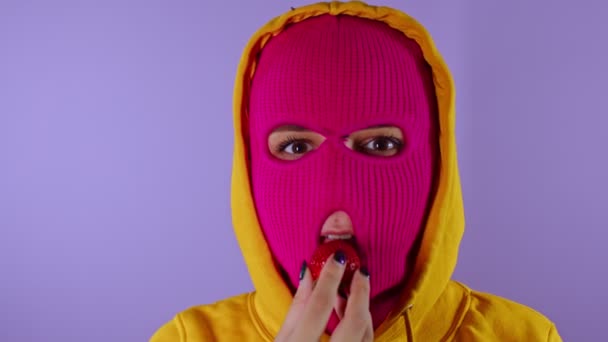 Close up of young woman in pink balaclava and yellow hood eats strawberry on purple background. Unknown female in mask bites ripe berry, looking at camera. — Stock Video