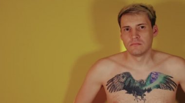 Göğsünde dövme olan çıplak vücutlu yakışıklı bir adam sarı arka planda duran kameraya bakıyor.