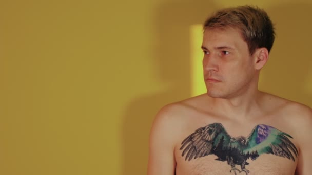 Egy jóképű férfi, csupasz törzzsel, tetoválással a mellkasán, félrenéz, sárga alapon áll. — Stock videók