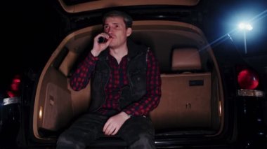 Genç adam geceleri otoparkta arabanın bagajında oturuyor. Düşünceli adam, bagajı açık bir şekilde arabasında otururken kafasını çevirir ve kaçar.