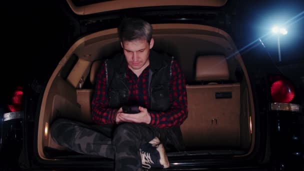 一个拿着手机坐在后备箱里的年轻人在晚上停车 一个成年人一边浏览智能手机一边坐在车里打开行李箱 — 图库视频影像