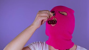 Pembe kar maskeli genç bir kadın mor arka planda tatlı kiraz yiyor. Maskeli meçhul kadın olgun meyvelerden ve gülümsemelerden hoşlanıyor.