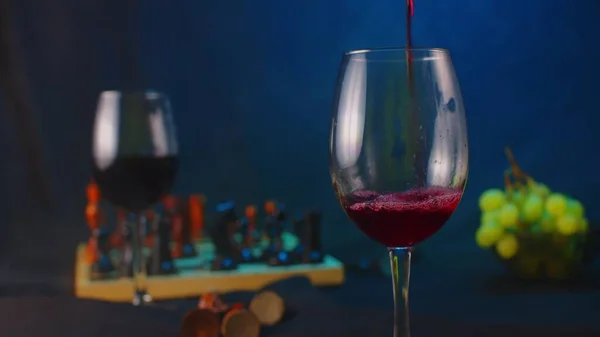 Still-life of empty glass in foreground and glass with red wine, board game, grapes in background. Close up of unrecognizable person pours red wine into empty glass