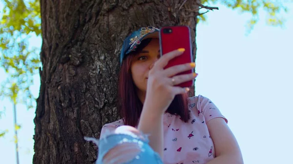 若い女性は携帯電話で 通りの木の近くに座っている キャップブラウジングスマートフォンのブルネット 夏に大きな木の近くの日陰に座って — ストック写真