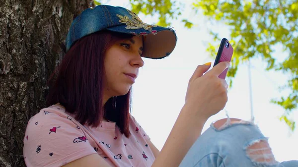 年轻的女人拿着手机 坐在树上的大街上 夏天的时候 戴着帽子的布鲁内特会浏览智能手机 坐在大树旁的树阴下 — 图库照片