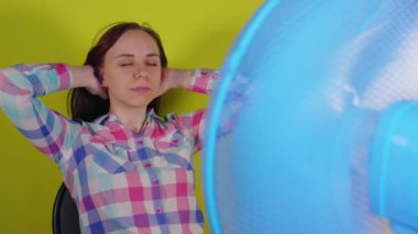 Sarı arka planda pervanenin önünde oturan genç bayana yakın çekim. Elleri başının arkasında mutlu bir esmer, solunum cihazının önünde otururken sıcaktan kaçıyor.