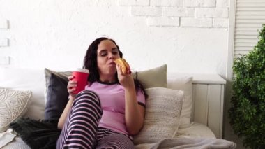 A young woman in bed eating a hot dog and drinking coffee. Breakfast in bed. Food delivery to your home.