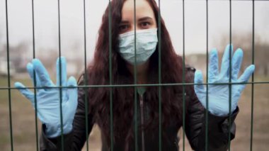 Yüzünde tıbbi maske olan genç bir kadının çitin arkasından yakın çekimi. Yetişkin bir kadın, yürürken kendini hastalıklardan korumak için yüzünü maskeyle örttü..