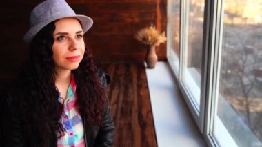 Şapkalı ve deri ceketli güzel bir kadın pencerenin yanında oturuyor. Gündüz vakti pencereden dışarı bakan güzel bir kadının portresi.