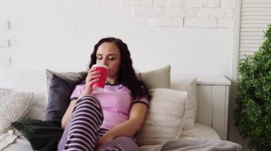 A young woman in bed eating a hot dog and drinking coffee. Breakfast in bed. Food delivery to your home. 