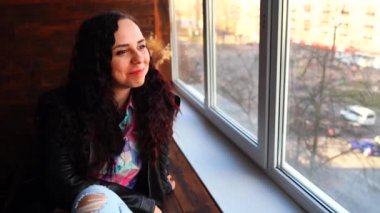 Deri ceketli genç ve güzel bir kadın pencerenin kenarında oturuyor. Güpegündüz kameraya bakan gülümseyen kadın portresi.