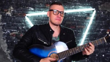 Aydınlanmış duvarın yanında gitar çalan dövmeli adam. Göğsünde kuş dövmesi olan şık bir adam gözlerini kaçırıp gitar çalarken eski püskü tuğla duvarın karşısında üçgen ışıklandırmayla dikiliyor.