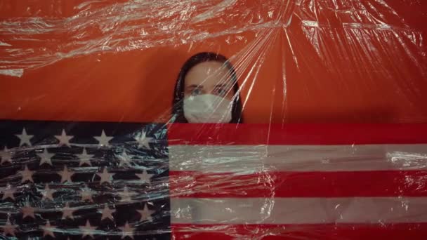 オレンジ色の背景にコロノウイルスから隔離されたアメリカ国旗を持つ医療用マスクの若い女性 病気から身を守るために仮面で顔を覆う女性の肖像 — ストック動画