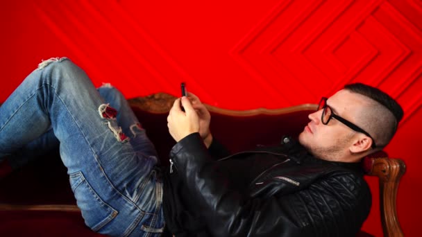 Joven Guapo Con Gafas Descansando Pequeño Sofá Contra Pared Roja — Vídeos de Stock