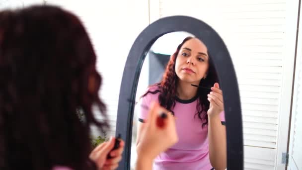 穿着睡衣的年轻漂亮的女人用睫毛膏画上了她的睫毛 在镜子里看着家里 在大镜子前把迷人的女性打扮得漂亮 — 图库视频影像
