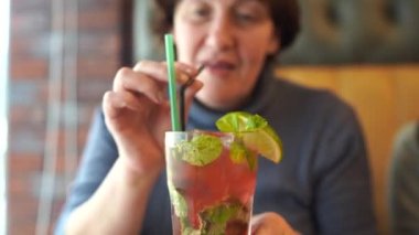Yetişkin bir kadın kafede kokteyl içiyor. Yetişkin dişi, masada otururken samandan taze meyve içeceği yudumluyor.