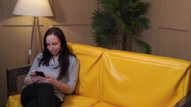 Evdeki kanepede dinlenen kendinden emin genç bir bayan. Evdeki sarı kanepede akıllı telefonuyla otururken kameraya bakan gündelik giysiler içindeki neşeli, genç bir kadından.