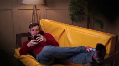 Sarı kanepede uzanmış gündelik giysili genç adam. Yetişkin erkek dinleniyor ve çeşitli haberlere cep telefonuyla bakıyor..
