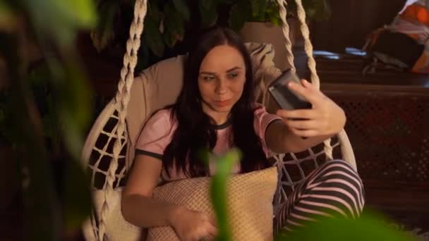 快乐的年轻女人坐在吊床上 坐在家里拿着智能手机 快乐的年轻貌美的女性坐在家里的吊床上 一边看着镜头一边看着镜头 — 图库视频影像