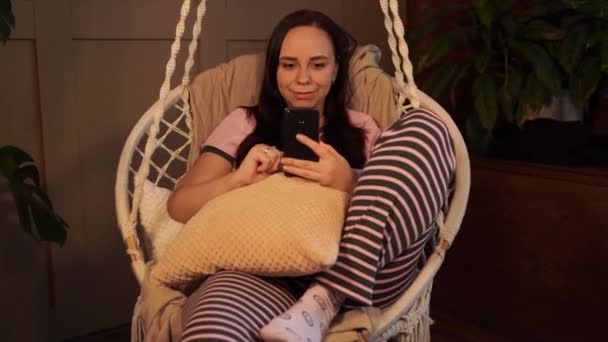 幸せな若い女性ハンモックに自宅でスマートフォンで座って休んでいる 陽気若いです魅力的な女性見ますカメラで座っている間にクッションでハンモック椅子にホーム — ストック動画