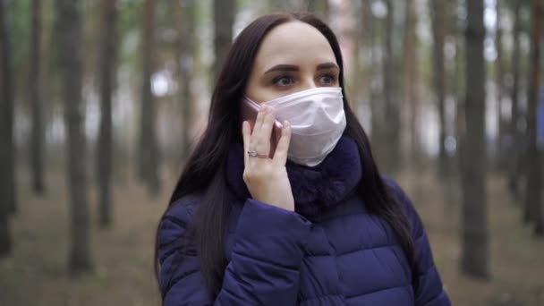 森の中を歩いている彼女の顔に医療マスクの若い女性と携帯電話の流行についてのニュースを学ぶ 大人の女性は病気から身を守るためにマスクで顔を覆いました — ストック動画