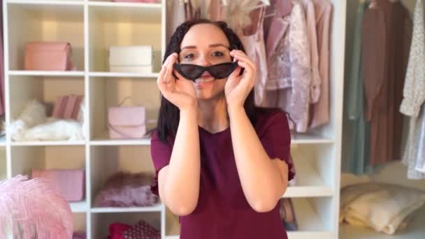 快乐的年轻女人在更衣室里试戴太阳镜 看着站在衣柜里戴着黑色太阳镜的镜头 快乐而迷人的女性 — 图库视频影像