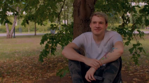 年轻人的近身休息 坐在公园的树上 他坐在公园的一棵树旁 一边听着 一边高兴地听着 — 图库视频影像