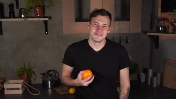 若い男が台所に果物を投げます 大人ハンサムな男イチャイチャと見ますのオレンジでカメラ — ストック動画