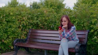 Cep telefonlu genç bir kadın, çalılarla çevrili bir bankta oturuyor. Kadın akıllı telefon, Parkland 'da bankta oturuyor..