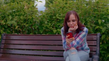 Cep telefonlu genç bir kadın, çalılarla çevrili bir bankta oturuyor. Kadın akıllı telefon, Parkland 'da bankta oturuyor..