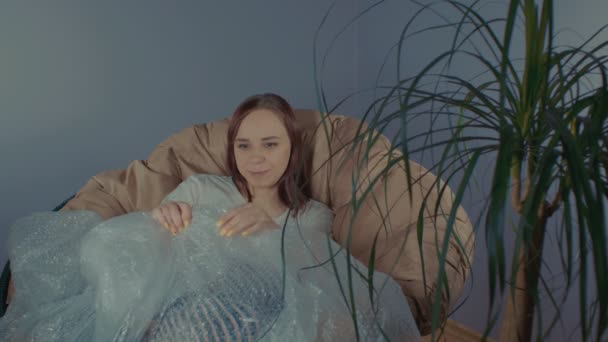Młoda Kobieta Bawi Się Folią Bąbelkową Siedząc Fotelu Pokoju Brunetka — Wideo stockowe
