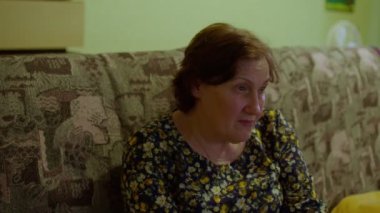 Olgun bir kadının kanepede oturup odada televizyon izlediği yakın plan. Düşünceli yaşlı kadın kanepede dinleniyor ve en sevdiği programı izliyor..