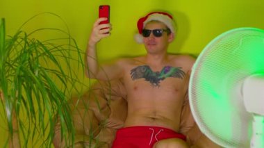 Noel Baba şapkalı, çıplak vücutlu genç adam akıllı telefondan selfie çekiyor, yelpazeden önce koltukta oturuyor. Güneş gözlüklü yetişkin adam tatil yerindeki tatilinin tadını çıkarıyor. İzolasyon kavramı.
