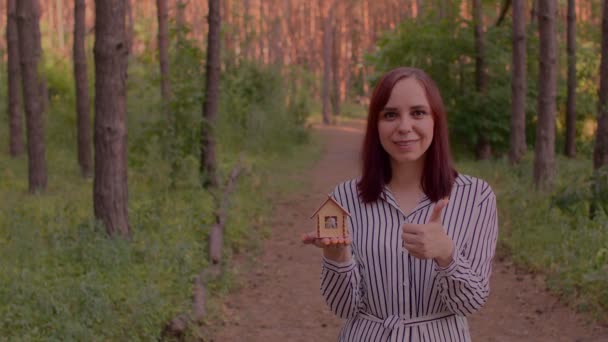 若い女性は小さな木造の家を保持し 森の中で親指を表示します 新しいアパートや屋根のオーバーヘッドを購入する概念 — ストック動画