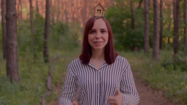 森の中に親指を示す頭の上に小さな木造の家を持つ若い女性 新しいアパートや屋根のオーバーヘッドを購入する概念 — ストック動画