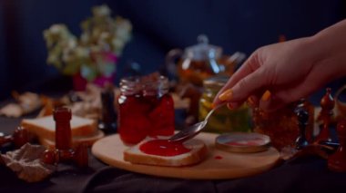 Reçelli küçük kavanoz ve tahta tahtada bir parça ekmek. Tanınmayan bir kadın çay kaşığıyla tostun üzerine reçel sürüyor..