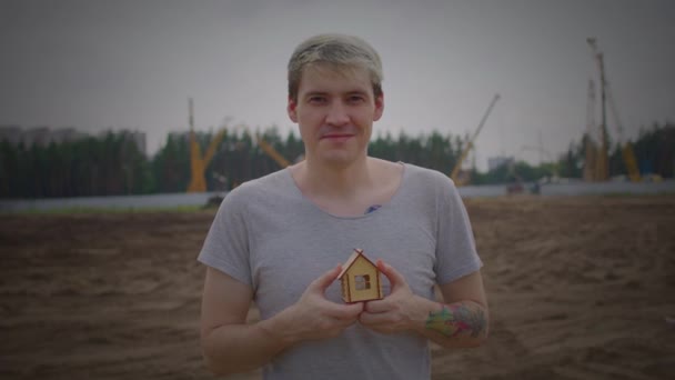 手に小さな木造の家を持つ若い男が建設現場に立っています。新しいアパートや屋根のオーバーヘッドを購入する概念 — ストック動画