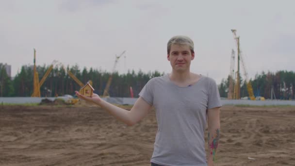 Joven con una pequeña casa de madera en la mano se encuentra en el sitio de construcción. Concepto de compra de apartamento nuevo y techo. — Vídeo de stock