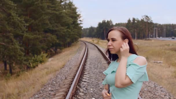 Figyelmes nő zenét hallgat, a vasúton áll vidéken. Fiatal nő fülhallgatóval, félrenéz a természetben. A veszélyes vasúti helyzet fogalma. — Stock videók