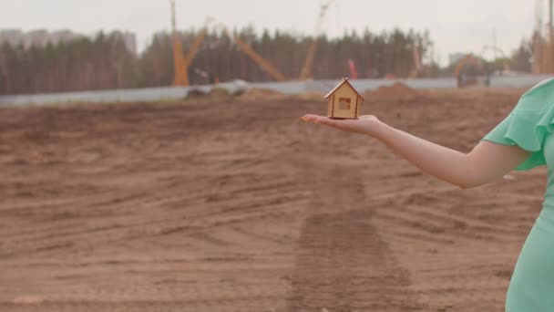 Mujer joven con una pequeña casa de madera en la mano se encuentra en el sitio de construcción. Concepto de compra de apartamento nuevo y techo. — Vídeos de Stock