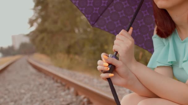 Крупним планом частина тіла жінки з парасолькою, що сидить на залізниці в похмуру і вітряну погоду. Нерозпізнана людина чекає дощу, сидячи на рейці — стокове відео