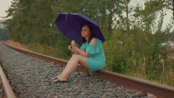 Junge Frau mit Regenschirm sitzt bei trübem und windigem Wetter auf der Bahn. Erwachsene Frauen warten auf Regen, sitzen auf Schienen und schauen in der Natur weg. — Stockvideo