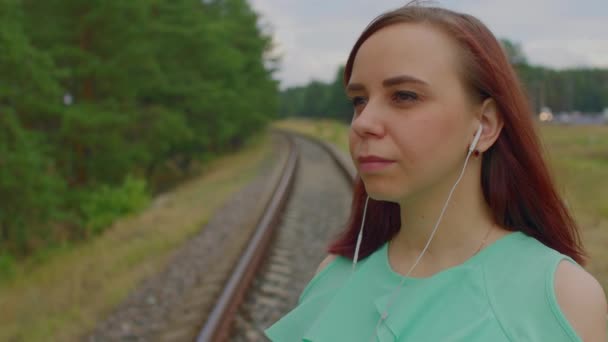 Femme réfléchie écoute de la musique, debout sur le chemin de fer dans la campagne. Jeune femme avec écouteurs regardant loin dans la nature. Concept de situation dangereuse sur le chemin de fer. — Video