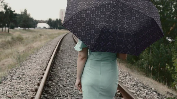 Rear view of young woman with umbrella standing on railway. Unrecognizable female with umbrella standing on railroad in cloudy weather