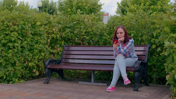 전화를 가지고 여자는 관목으로 둘러싸인 벤치에 공원에서 벤치에 스마트폰을 — 스톡 사진