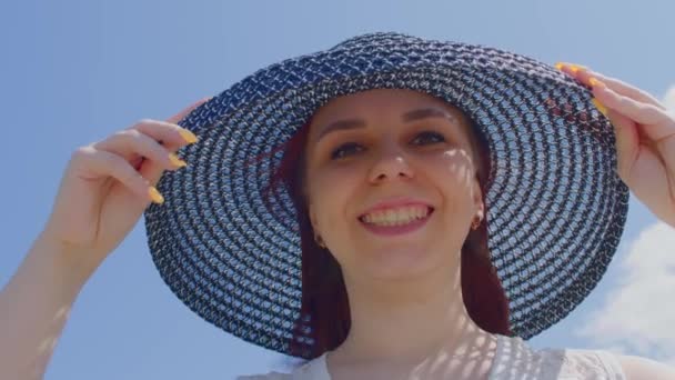 身穿白衣 头戴雅致帽子的年轻女子站在丘陵地带 美丽的女人走出帽子 在阳光灿烂 多风的天气里看着相机 — 图库视频影像