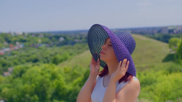 在多山的天气里 年轻的女性穿着白衣 戴着雅致的帽子 阳光灿烂 美丽的女人紧紧抓住她的帽子 站在郊外的室外 — 图库视频影像
