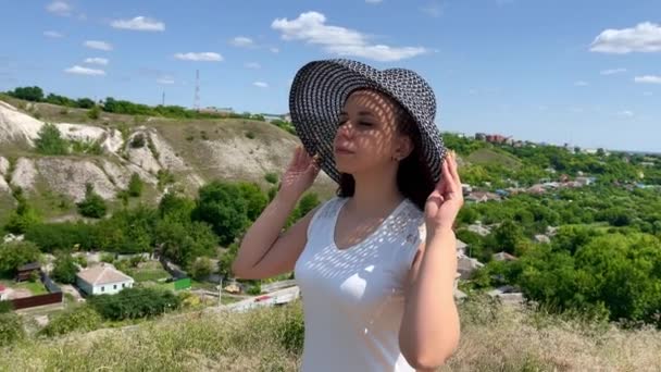 Mujer joven en vestido blanco y sombrero elegante en día soleado en terreno montañoso. Hermosa mujer con los ojos cerrados se aferra a su sombrero, de pie al aire libre en el campo. — Vídeo de stock